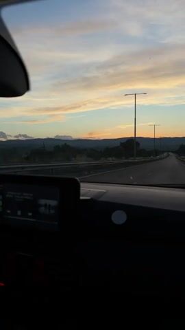 The video shows a first-person view from inside a car driving on a highway at dusk. The sky is painted with hues of orange, pink, and blue as the sun sets behind a distant mountain range. Tall streetlights line the highway, illuminating the road ahead. The car's dashboard is visible in the foreground, with a digital screen displaying what appears to be music information. Trees and foliage line the sides of the road, creating a scenic backdrop for the journey. The overall atmosphere is serene and tranquil, suggesting a peaceful drive as day transitions into night.