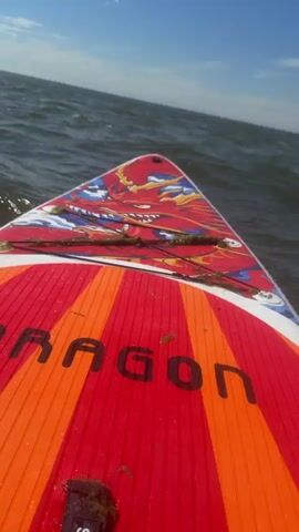 The video shows a first-person perspective from a paddleboard on the water. The paddleboard is red and orange with the word "DRAGON" visible. There are some pieces of seaweed on the front of the board. The water is choppy with small waves, and the sky is blue with some white clouds. In the second and third clips, a red paddle with a black shaft is visible being used to propel the board through the water. The overall scene depicts a person paddleboarding on a sunny day.