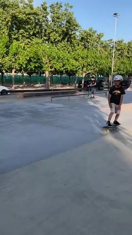 The video features two males skateboarding at an outdoor concrete skate park in Santa Perpètua de Mogoda, Spain, during what appears to be a sunny late afternoon or early evening.

The primary subject is a younger male, wearing a white helmet with visible text "CHIN", a black t-shirt with a graphic, and light-colored shorts. He is skateboarding across a flat concrete area, then approaches a low metal rail. He attempts a trick, placing his skateboard perpendicularly onto the rail as if to grind or slide, before stepping off and returning to his board on the ground.

Another male, appearing older, wearing a green t-shirt and grey pants, is also skateboarding in the background throughout the video, further back in the skate park.

The setting includes various skate park features like ramps and flat ground, surrounded by numerous green trees. In the background, a road with cars driving by and a white fence are visible, along with a bridge in the distance in some frames. The sky is clear and blue.