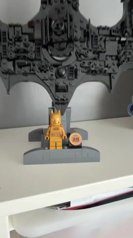 The video shows a black LEGO Batman logo and a gold Batman figurine on a white shelf. A hand reaches out and opens a hinged section of the logo to reveal a black Batman figurine. The shelf has white bins below it.