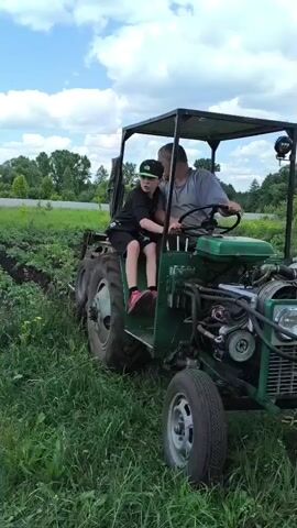 A middle-aged man drives a green tractor with a canopy on a grassy field. A boy sits next to him. The tractor has a visible engine and two headlights. The field appears to be a garden or farm, with rows of plants visible. Trees line the background, and the sky is cloudy.