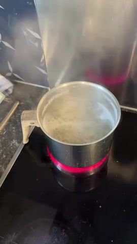 The video begins by showing a metal pot filled with water boiling vigorously on a black induction hob, which glows red beneath the pot. Steam is visibly rising from the boiling water. The camera then pans quickly to the right, revealing a kitchen counter.

The view settles on a white plate where two halves of a burger bun are placed. One bun half is topped with melted orange cheese, while the other toasted half appears buttered. A silver knife rests on the plate, next to a black slotted spoon holding a few pieces of cooked pasta. In the background, a silver toaster is visible on the kitchen counter, along with a tablet device to the left of the plate. The scene depicts a kitchen setting during meal preparation. No people are visible. No discernible text is present.