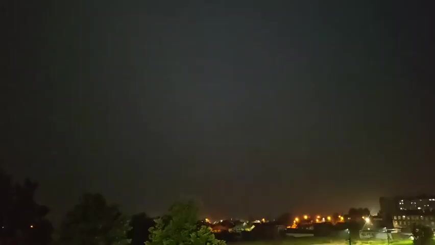 The video captures a nighttime scene over a rural or suburban landscape. The sky is predominantly dark, with a faint purple hue, indicating twilight or overcast conditions. Scattered lights from buildings and streetlights are visible in the distance, suggesting a small town or village. Trees frame the lower left and center of the image, their leaves dark against the night sky. The overall atmosphere is calm and still, with no discernible activity or movement other than a very subtle change in the sky's color and brightness.