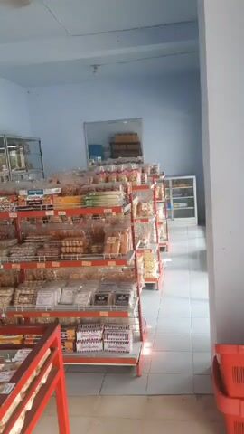 The video shows the interior of a small shop, possibly a convenience store or a bakery. There are several shelves filled with packaged goods, including snacks, cookies, and biscuits. The shelves are made of metal with red framing. In the background, there is a mirror on the wall and some storage boxes stacked behind the shelves. The lighting appears to be artificial. There are no people visible in the video. The floor is tiled.