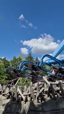 A short video clip shows a roller coaster ride with multiple cars filled with passengers. The cars are blue and black and are moving along a track. The passengers are seated and appear to be enjoying the ride. The background shows trees and a blue sky with white clouds. The roller coaster cars are attached to a blue support structure. The foreground shows a rocky area with wooden fencing. The roller coaster appears to be moving uphill.