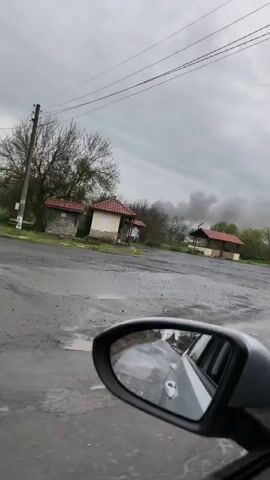 The video is filmed from inside a moving vehicle, with a portion of the car's side mirror visible in the lower right foreground, reflecting the car's exterior. The scene depicts a wide, open area with a ground surface that appears to be a mix of dirt and gravel, possibly wet. The sky is heavily overcast with thick grey clouds.

In the mid-ground, several small, single-story buildings with reddish-brown tiled roofs are visible. On the left, two simple structures stand close together, while further to the right, another open-sided structure, resembling a gazebo or shelter, is seen. Bare trees, characteristic of late winter or early spring, are on the left side of the frame, with some distant trees showing green foliage.

A prominent feature in the background is a large, dark plume of smoke rising against the grey sky, indicating a fire or significant burning activity in the distance. As the vehicle capturing the video moves, a blue car towing a white tanker trailer drives across the background from left to right. The overall impression is of a rural or semi-rural landscape on a cloudy day in Livada, Bulgaria, with a noticeable smoke plume in the distance.