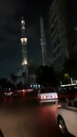 The video shows a mosque with two tall minarets, illuminated at night.  Cars are driving on the road in front of the mosque, their headlights and taillights visible. A tall building is seen on the right side of the frame. The sky is dark and cloudy.