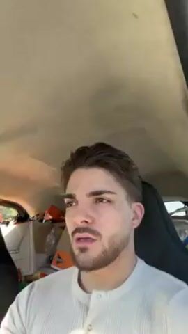 The video shows a close-up view of a young man with short, dark brown hair and a trimmed beard. He is wearing a light-colored, button-down shirt. The man is looking off to his left, with a slightly concerned or contemplative expression. The setting appears to be inside a car, with the light grey ceiling visible above him. The back of the car is filled with boxes and bags, including what appears to be a Cheetos box. The car seat is black. The camera is relatively still, with only slight movements.