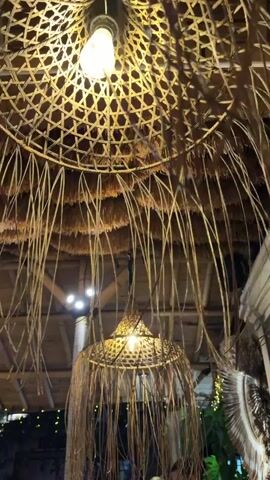 The video shows an interior view looking up at two thatched light fixtures with Edison-style bulbs. The light fixtures are hanging from a thatched ceiling. The upper fixture is larger and more intricately woven with thin, dry grass strands hanging from it. The lower fixture is smaller and also made of woven grass, with similar strands hanging down. The background shows the wooden structure of the ceiling and some distant lights.
