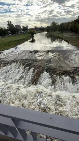 The video shows a river flowing over a small dam, creating a waterfall effect. The view is from a bridge with gray railings. The water is turbulent and foamy at the base of the dam. Upstream, the river is calmer, reflecting the sky. The sky is cloudy.  The riverbanks are grassy with trees lining them.  In the distance, buildings can be seen.