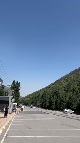 A vertical video pans across a scenic mountain landscape under a clear blue sky. The scene begins in a paved parking lot with white lines marking spaces. A white SUV is parked in one of the spots, and a silver car drives past on the road adjacent to the parking lot. A dense forest of evergreen trees covers the steep slope of a mountain that dominates the right side of the frame. Further into the video, a small building with a sign that reads "БАЙЧЕЧЕКЕЙ" appears, followed by a modern building with a "WC" sign. In the distance, cable cars can be seen ascending the mountain, and snow-capped peaks are visible. The video concludes with a view of a larger building with a sign that appears to be a ski resort or mountain lodge, with several vehicles and people milling about in front of it. The overall impression is of a well-developed tourist area in a beautiful mountain setting.