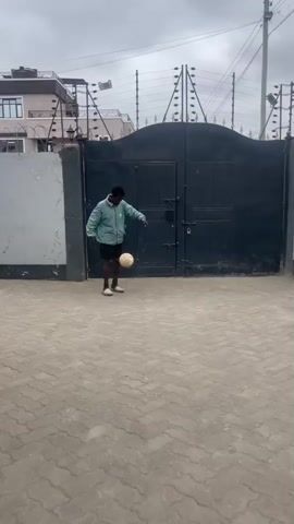 A young Black male is standing on a paved driveway in front of a large, dark metal gate. He is wearing a light blue jacket, black shorts, and white sneakers. He is juggling a soccer ball with his feet, keeping it in the air. The gate is closed and appears to be electrified with security wiring along the top. To the left of the gate is a portion of a building, and to the right, a utility pole. The sky is overcast.