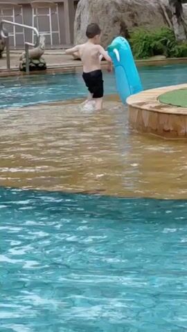 A young boy walks in a shallow pool near an inflatable blue seal. A palm tree stands in the middle of the pool. A woman holds a baby in the water. A young girl stands in the water with her arm outstretched.