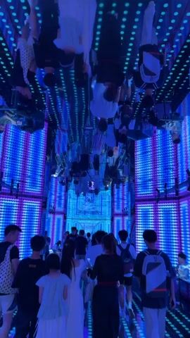 The video shows a group of people walking through a long, narrow hallway. The hallway is lined with LED lights that are blue and purple. The ceiling is also lined with lights, and there are mirrors on the ceiling that reflect the lights and the people in the hallway. The people in the video are mostly young adults and teenagers, and they are dressed in casual clothing. Some of the people are taking pictures or videos with their phones. The overall atmosphere is festive and exciting.