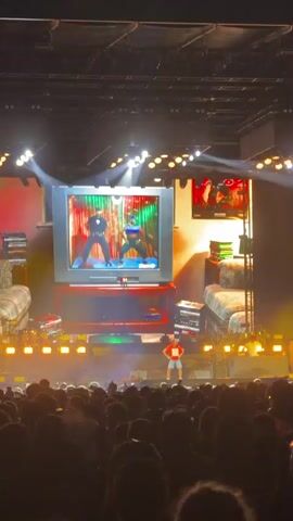 A stage performance is captured from a low angle, looking up at a large screen displaying two people in athletic wear, seemingly participating in a fitness video game. The screen is set within a mock living room environment, complete with two sofas, stacks of what appear to be video game cases, and a vintage television set. On the stage in front of the screen, a single person in a red shirt and shorts is facing the audience. The stage is illuminated by bright yellow stage lights, with additional spotlights creating dramatic effects. The audience is visible in the foreground, largely obscured by darkness. The overall impression is a retro-themed entertainment show.