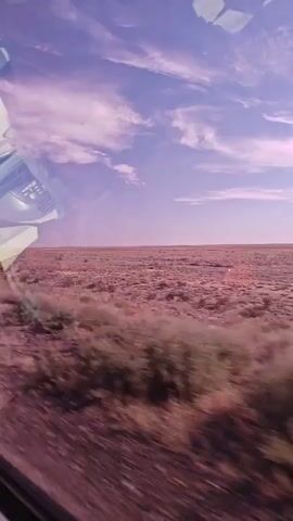 The video shows a landscape from a moving vehicle. The sky is a purplish-blue with scattered clouds. Below the sky is a vast, flat expanse of reddish-brown land covered with low-lying vegetation. In the foreground, the ground appears to be dirt and gravel, with some green brush along the side of the road. There is a blurred object, possibly part of the vehicle or another structure, visible in the upper left corner of the frame. The overall impression is that of traveling through a semi-arid or desert environment.