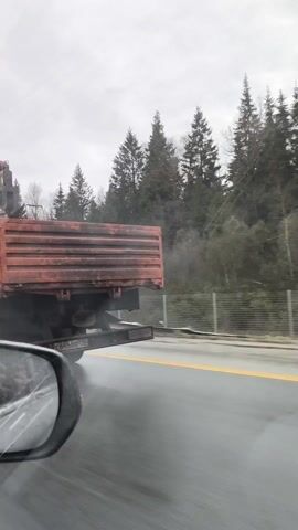 The video shows a yellow and red truck driving on a highway. The truck has an orange bed with a red crane attached. The road is wet and has a yellow dividing line. The weather is overcast, and there are trees visible in the background. The view is from the side of a car, showing the side mirror. The truck is identified as a PALFINGER INMAN IT 150.