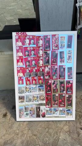 A flat lay display of merchandise is shown. The display is white and rectangular, and it is placed on a surface that appears to be a sidewalk or pavement. The merchandise consists of numerous rectangular and square items, predominantly featuring images of Joseph Stalin and Soviet propaganda symbols like the hammer and sickle, set against red backgrounds. There are also images of pomegranates and scenes of Georgian architecture and food. Some items have text in what appears to be Georgian script. The price "10 GEL" is visible at the top of the display. The lighting suggests an outdoor setting, possibly in daylight. No people are visible in the frame, and no actions are being performed.