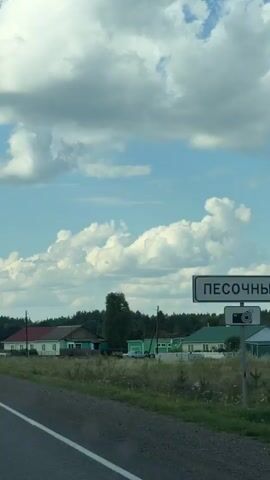 The video begins by showing a rural road with houses on the left side of the frame. The sky is blue with large, fluffy white clouds. As the video progresses, the perspective shifts to a view from inside a vehicle driving on a paved road. The road is bordered by a grassy shoulder and trees. Streetlights are visible along the side of the road, and there are occasional signs. The scenery is predominantly natural, with rolling hills and dense vegetation. The overall impression is a drive through a scenic, possibly rural, landscape under a bright, cloudy sky.