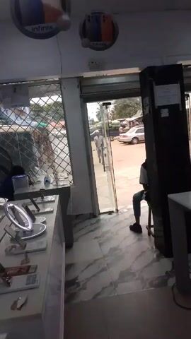 The video shows the interior of a mobile phone shop. There is a display counter with several smartphones and accessories on it. A fan is running near the window, which has a metal grill. An open doorway leads to a street with a car parked. A person is sitting on a stool near the doorway, facing outwards. Another person in a white shirt is entering the store through the open door. There are Infinix advertisement signs hanging from the ceiling. The floor is covered with marble-patterned tiles.