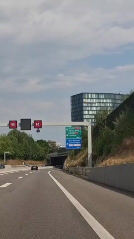 The video shows a view from a vehicle driving on a highway. Overhead signs indicate speed limits of 80 and provide directions to Zurich, Basel, Lausanne, Bern-Wankdorf, and Ostermundigen. A modern, glass-fronted building is visible on the right, behind a sound barrier and vegetation. The sky is cloudy.
