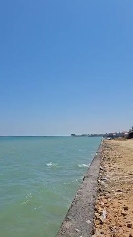The video shows a serene view of the sea under a clear blue sky. The water has a greenish-blue hue and gentle waves lap against a rocky shore in the foreground. A person is sitting on the rocks, facing the sea, their back to the camera. In the distance, a wooden pier extends into the water, with several small structures on it. The overall impression is one of tranquility and natural beauty.