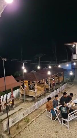 The video shows an outdoor dining area at night. There are several bamboo huts with thatched roofs, each with people sitting inside. A white picket fence runs along the edge of the seating area. In the foreground, there are white picnic tables with people dining. A two-story building with an open-air second floor is visible in the background, lit by string lights. There is a staircase leading up to the second floor. The ground is covered in gravel. Several motorcycles are parked to the right of the building. The overall atmosphere is casual and inviting.