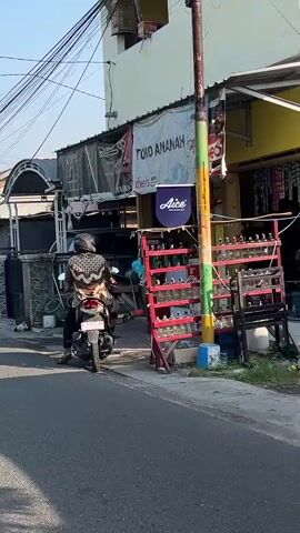 A person wearing a patterned shirt and helmet is sitting on a motorcycle parked on the side of a street. The motorcycle is facing away from the camera. To the right of the motorcycle is a building with a sign that reads "TOKO AMANAH." In front of the building are shelves filled with bottles, possibly fuel. A red barrel is also visible. The street appears to be paved, and there is a sidewalk. The lighting suggests it is daytime.