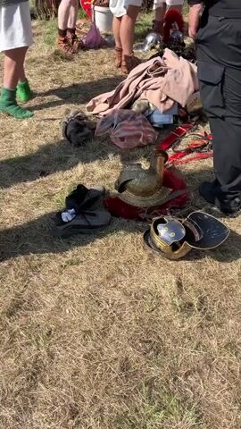 This video captures an outdoor scene featuring several men, primarily dressed in Roman-style military reenactment attire, gathered on a dry, grassy field. The bright daylight casts distinct shadows, indicating a sunny day.

In the foreground and midground, multiple individuals are visible. One man on the left wears a chainmail tunic (lorica hamata) over a white undergarment, paired with bright green socks and sandals. Another man, seen from the back, is dressed in a red tunic with a leather belt. A third prominent figure wears segmented plate armor (lorica segmentata) over a white tunic, appearing to adjust his gear. Other reenactors in similar Roman-era clothing and armor are partially visible in the background and around the central group. One man on the far right is dressed in modern black tactical pants and a dark shirt, observing the activity.

Scattered on the ground are various items, including elements of Roman military equipment and personal belongings. Two ornate Roman helmets (galea) are clearly visible: one brass-colored with intricate detailing and another silver-colored, both featuring red crests or plumes. Piles of fabric, likely tunics or cloaks, lie on the dry grass. Modern items such as black sandals and shoes are also mixed in with the historical gear.

The scene suggests a historical reenactment event, possibly either preparations before a demonstration or the packing up of equipment afterward. The background shows additional people and tents or structures, indicating a larger public gathering or festival. There is no visible text in the frame.