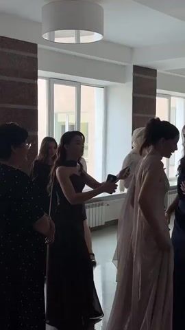 In a brightly lit indoor hallway, a group of young women in formal attire appears to be gathering. The hallway features large windows and beige tiled walls. In the foreground, a woman in a dark, strapless gown with a high slit is holding two phones and looking down. To her left, another woman in a similar dark gown stands facing away. Further back, a woman in a white hijab and dress is holding a phone. To the right of the woman in the hijab, a woman in a dark blue gown is looking away, and the back of a woman in a light-colored gown is visible. In the background, a large banner displaying the year 2026 and various words in Russian suggests a celebratory event, possibly a graduation or prom. White decorative trees are also present. The women are dressed in floor-length gowns, indicating a formal occasion.