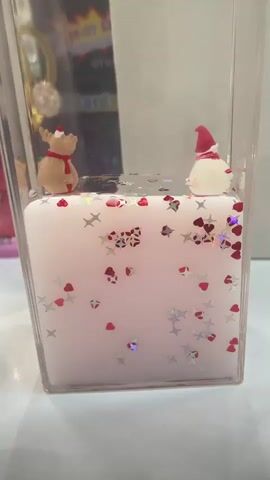 A close-up, eye-level shot shows a clear plastic container with a white candle inside. The candle is decorated with red and silver glitter in the shape of hearts and stars. Two small figurines, a reindeer and a snowman wearing Santa hats, are placed on top of the candle. The background is blurred and appears to be a shelf with various objects, including a bright, colorful poster. A human finger is visible on the left side of the frame, holding the container. The lighting is bright, suggesting an indoor setting. The video captures subtle movements of the glitter within the container, likely due to the gentle handling of the object.