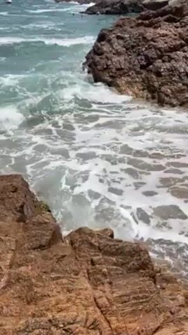 Certainly! Here's a detailed description of the video:

**Scene:** The video captures a coastal scene with waves crashing onto a small beach nestled between rocky formations. 

**Environment:**
*   **Rocks:** The foreground is dominated by large, reddish-brown rocks. Their texture suggests they've been shaped by the sea over time.
*   **Beach:** A small, sandy beach is visible, partially submerged as waves roll in.
*   **Ocean:** The water is a mix of turquoise and white where the waves are frothing. The horizon is visible, suggesting an open ocean beyond.

**Action:**
*   **Waves:** The primary action is the constant crashing of waves against the rocks and beach. The foamy, white water indicates the force of the waves.

**Composition:**
The camera is positioned high up, providing an overview of the cove. The rocks and beach are in the foreground, with the ocean stretching into the background.

Overall, the video conveys a sense of the rugged natural beauty of a rocky coastline, with the waves being the dynamic and dominant force in the scene.