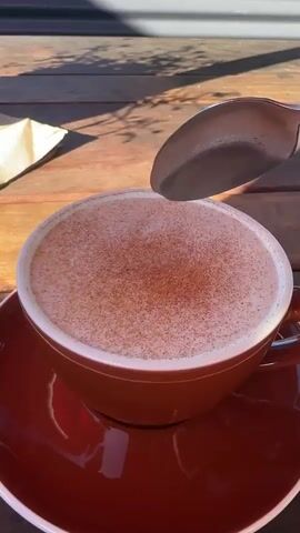 A close-up shot shows a brown mug filled with a frothy drink, possibly hot chocolate or a latte, topped with a sprinkle of cinnamon or cocoa powder. A silver spoon is used to scoop a dollop of the foam from the mug. The mug and saucer are a deep red color and rest on a wooden surface, likely a table. Sunlight casts shadows on the table, suggesting an outdoor setting.