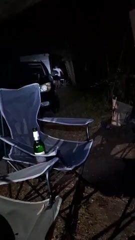 The video shows a dark outdoor scene at night. In the foreground, a folding camping chair is prominently displayed, with a bottle of green glass with a white label resting in its cup holder. Another folding chair is partially visible in the lower left corner. In the background, a dark-colored SUV is parked, its headlights illuminating the area. There appear to be people near the SUV, possibly gathered around a table or engaged in conversation, though they are not clearly visible due to the darkness and distance. The environment suggests a campsite or a similar outdoor gathering. The lighting is dim, with the headlights of the SUV and potentially a low-angle light source casting shadows.