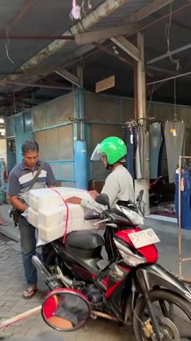 A man with graying hair and a striped shirt is standing next to a motorcycle. He is holding a large package wrapped in white material and secured with red straps. Another person wearing a bright green helmet and a light-colored shirt is seated on the motorcycle, reaching out to take the package. The motorcycle has a red seat and chrome accents. In the background, there appears to be a market stall with clothing racks and a blue structure. The ground is paved. The interaction seems to be a transfer of goods.