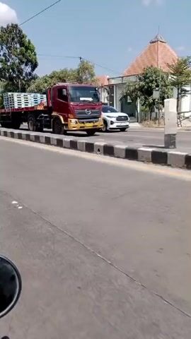 The video shows a view from a moving vehicle traveling on a paved road. The road has a dashed white line in the center and black and white striped barriers along the right side. Several trucks and cars are visible on the road. In the background, there is a large, ornate archway with the words "SELAMAT JALAN" written on it. Trees and buildings are also present. The sky is blue with some white clouds. The overall scene depicts a road leading out of a town or city, indicated by the archway which likely signifies an exit.