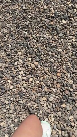 A first-person perspective video shows a person walking on a surface covered in small, multicolored pebbles. The camera is positioned to look down at the person's feet and legs. The person is wearing white athletic shoes with light blue and purple accents. The pebbles are predominantly shades of gray, brown, and tan, with some lighter-colored stones mixed in. The lighting suggests it is daytime and sunny, casting shadows on the pebble ground. The person's legs are visible from the knees down, and a tattoo is partially visible on the left calf. The movement is steady, indicating a slow to moderate walking pace. The video is brief, capturing only a few seconds of the walk.