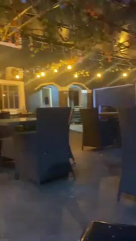The camera pans across an outdoor dining area at night. The area is decorated with hanging string lights and artificial vines. There are black wicker chairs and tables. A large standing fan is visible. The ground appears to be concrete. Music is playing in the background. The area appears empty.