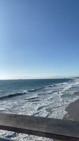 The video pans across a sunny beach scene. The camera begins by focusing on the ocean with gentle waves breaking on the shore. The sun is bright, casting a glare on the water. As the camera pans right, it reveals a sandy beach populated with a few people. In the background, there are buildings that appear to be houses or apartments lining the coast. There are also some beach amenities visible, such as volleyball nets and a lifeguard stand. The sky is clear and blue throughout the video.