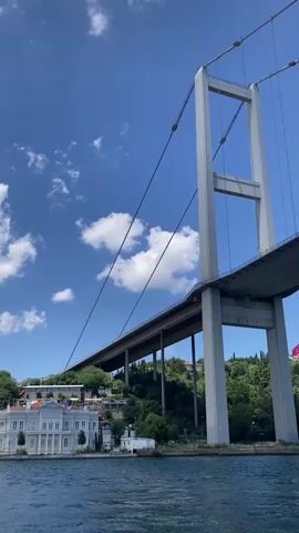 The video captures a low-angle shot of a large suspension bridge on a clear day with blue skies and scattered white clouds. A body of water, likely a river or strait, occupies the bottom portion of the frame, showing gentle ripples. The bridge's massive concrete support pillar is prominent on the right, with suspension cables extending upwards and outwards. Beneath the bridge deck, a sloping green hillside is visible, dotted with trees and some buildings. On the left side of the hillside, a white building with multiple windows and a distinct architectural style is partially visible. The overall impression is one of a grand man-made structure set against a natural landscape under a bright, sunny sky.