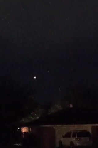 The video opens with a dark night sky filled with numerous small, faint stars. In the upper left quadrant, a single bright light, possibly a planet or a star, is visible. The scene transitions to show the silhouette of trees and the roof of a house with a car parked in front. For a brief moment, a cluster of bright lights appears in the sky, arranged in a scattered pattern. These lights seem to flicker and expand before the sky returns to its previous state, with only the faint stars and the single bright light in the upper left visible.