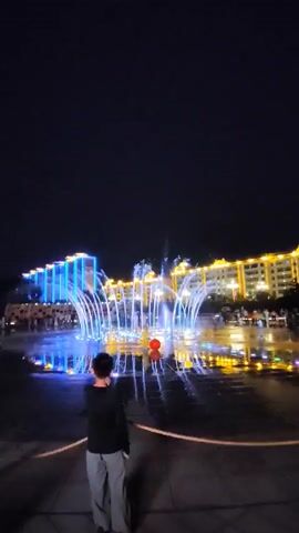 A vertical, nighttime video shows a person from behind standing in a plaza. The person is wearing a black long-sleeved shirt and gray pants. Before the person is a large, illuminated fountain with water jets spraying upwards in a circular pattern. The fountain is lit with blue and white lights. Behind the fountain are buildings that are also illuminated with blue and yellow lights. The ground in front of the fountain is wet, reflecting the lights from the fountain and buildings. There are other people in the background, but they are out of focus. The sky is dark.