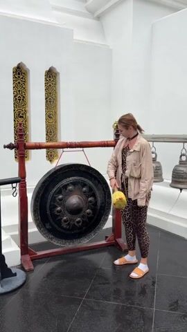 A woman stands in front of a large gong, holding a yellow mallet. She strikes the gong, producing a loud sound. The woman is wearing leopard-print leggings, a beige jacket, and orange slides. She is smiling and looking at the camera. In the background, there are several bells hanging from a rack. The scene appears to be outdoors, possibly at a temple or cultural site.