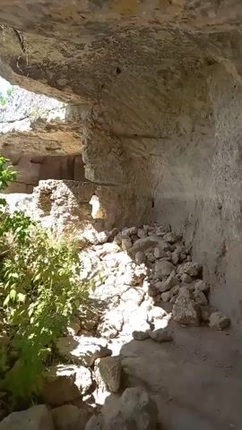 The video pans from inside a rocky overhang, showing crumbling stone walls and vegetation. It then moves to an opening that reveals a scenic view of a lush green forest and a blue sky with white clouds. The camera captures the natural environment, focusing on the textures of the rocks and the dense foliage.