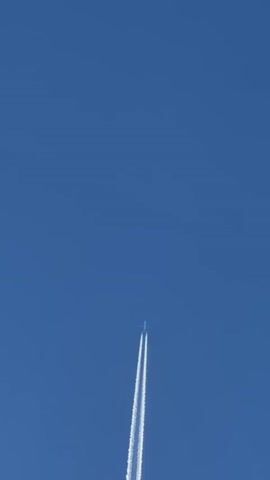 The video shows an airplane flying through a clear blue sky, leaving behind a white contrail. The airplane is a small white object at the top of the frame, and the contrail extends downwards, getting wider and more diffused as it moves away from the aircraft. The sky is a solid, uniform blue, with no clouds visible. The camera is positioned below the airplane, looking upwards. The video appears to be shot on a sunny day.