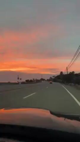 The video, shot from the driver's seat of a moving vehicle, captures a vibrant sunset. The sky is a mix of bright orange and dusky blue, with streaks of pink clouds. The vehicle is moving along a paved road with clear lane markings, and utility poles line the side of the road with visible power lines. Other vehicles are seen on the road ahead, and palm trees are visible on the right side of the road. The hood of the car is visible in the foreground, reflecting some of the orange sky.