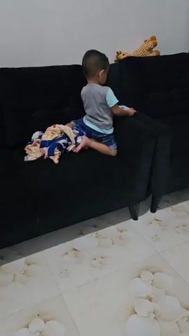 A young child sits on a dark sofa, facing away from the camera, with a phone in their hands. To the child's left, a colorful blanket is draped over the sofa. In the background, a stuffed giraffe toy rests on the sofa. The floor is tiled with a light-colored pattern. The child then turns to face the camera briefly before returning their attention to the phone. The video ends with the child looking down at the phone.