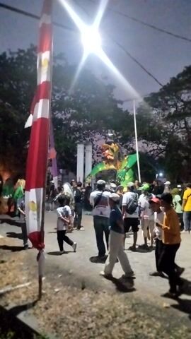 The video shows a group of people gathered around a large, colorful dragon effigy, likely during a parade or festival. The effigy is predominantly green with yellow and red accents, and its head is detailed with a fierce expression. A tall flagpole with red and white striped fabric is prominent in the foreground, and other festive decorations, including balloons, are visible. The scene appears to be outdoors, possibly at dusk or night, indicated by the dim lighting and artificial light sources. Some individuals are on motorcycles, suggesting a mobile procession. The people are dressed in casual attire, and their interactions suggest a lively and celebratory atmosphere. The camera angle is somewhat distorted and the focus is not always sharp, but the overall impression is one of a community event.