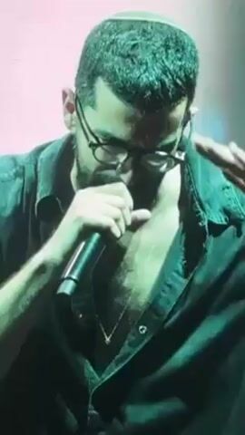 A bearded man with glasses and a yarmulke sings into a microphone. He wears an unbuttoned black shirt with a gold necklace visible. He moves his hand near his forehead as he sings, under green-tinged stage lighting.