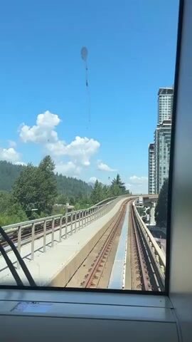 This is a POV shot from a train, looking out the front window. The train is on an elevated track, curving to the right. On the left, there is a walkway with a railing, and beyond that, trees and hills. On the right, there are tall buildings and more elevated tracks. The sky is blue with some white clouds. There is a light-colored mark on the window, possibly a smudge or a water spot, with a faint line extending down from it. The sun is shining brightly.